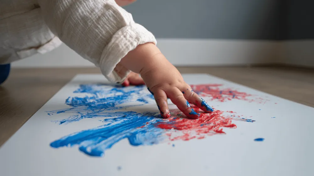 La mano de un niño pinta con los dedos en azul y rojo sobre un papel en el suelo.